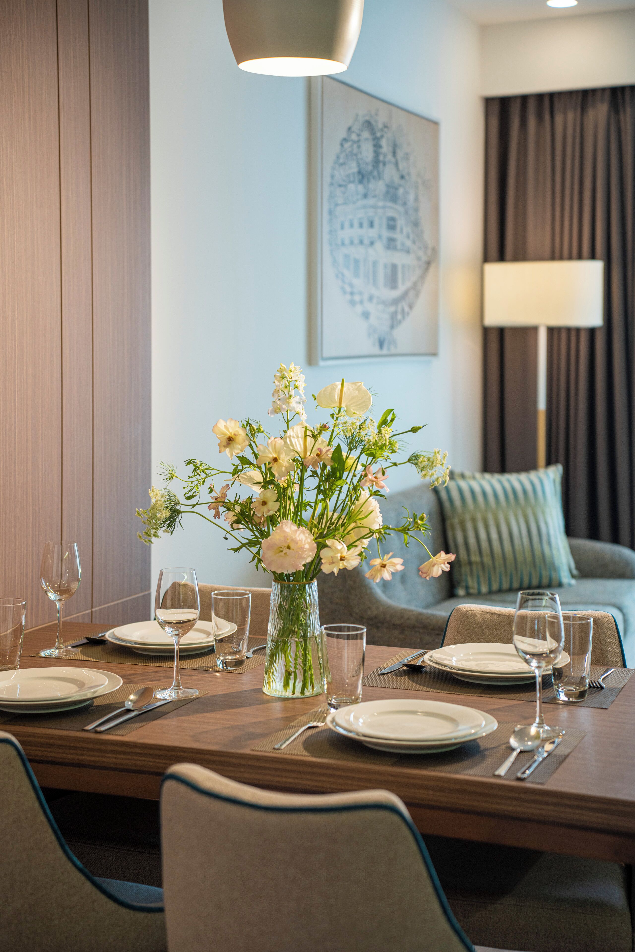A dining table set with plates, glasses, and cutlery, a vase with flowers, and a lamp in the background.