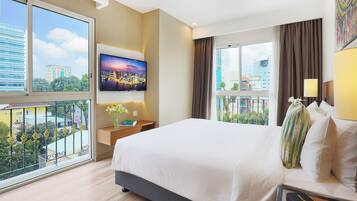 A hotel room with a large bed, a TV mounted on the wall, a balcony with a view of the city, and a nightstand with a lamp.