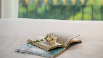 A bed with an open book and a flower, a window with a view of greenery and buildings, and a railing with decorative lights.