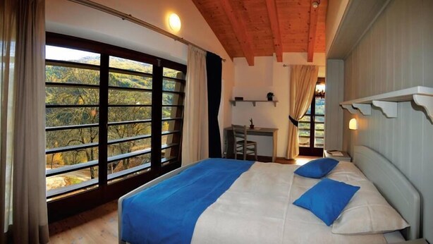 A bedroom with a large window, a bed with blue and white bedding, a desk, and a chair. - Agritur La Gemma (Sant'Orsola Terme)