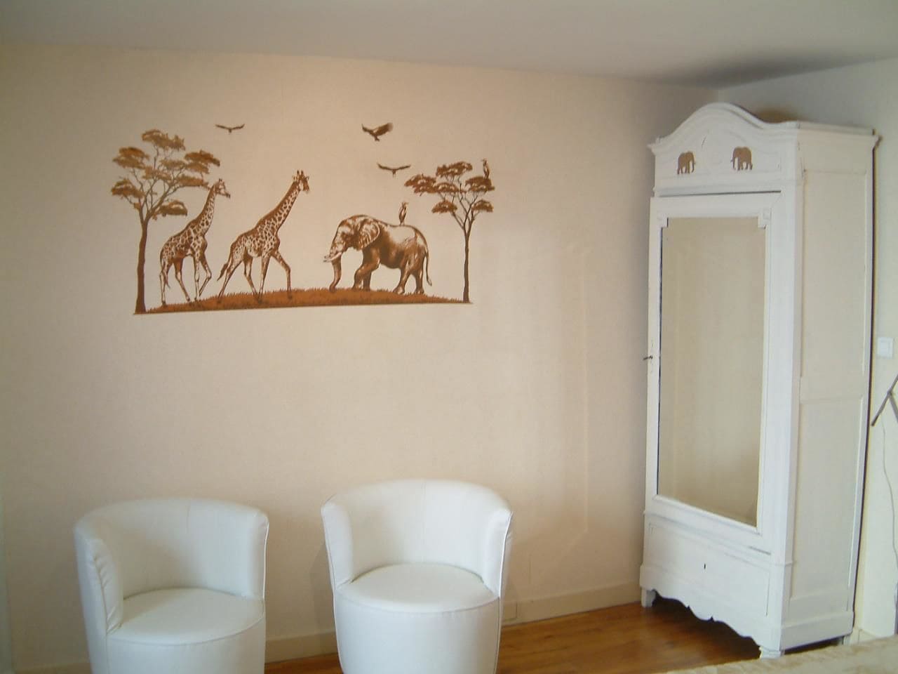A room with two white chairs, a white cabinet, and a wall mural of giraffes, elephants, trees, and birds.