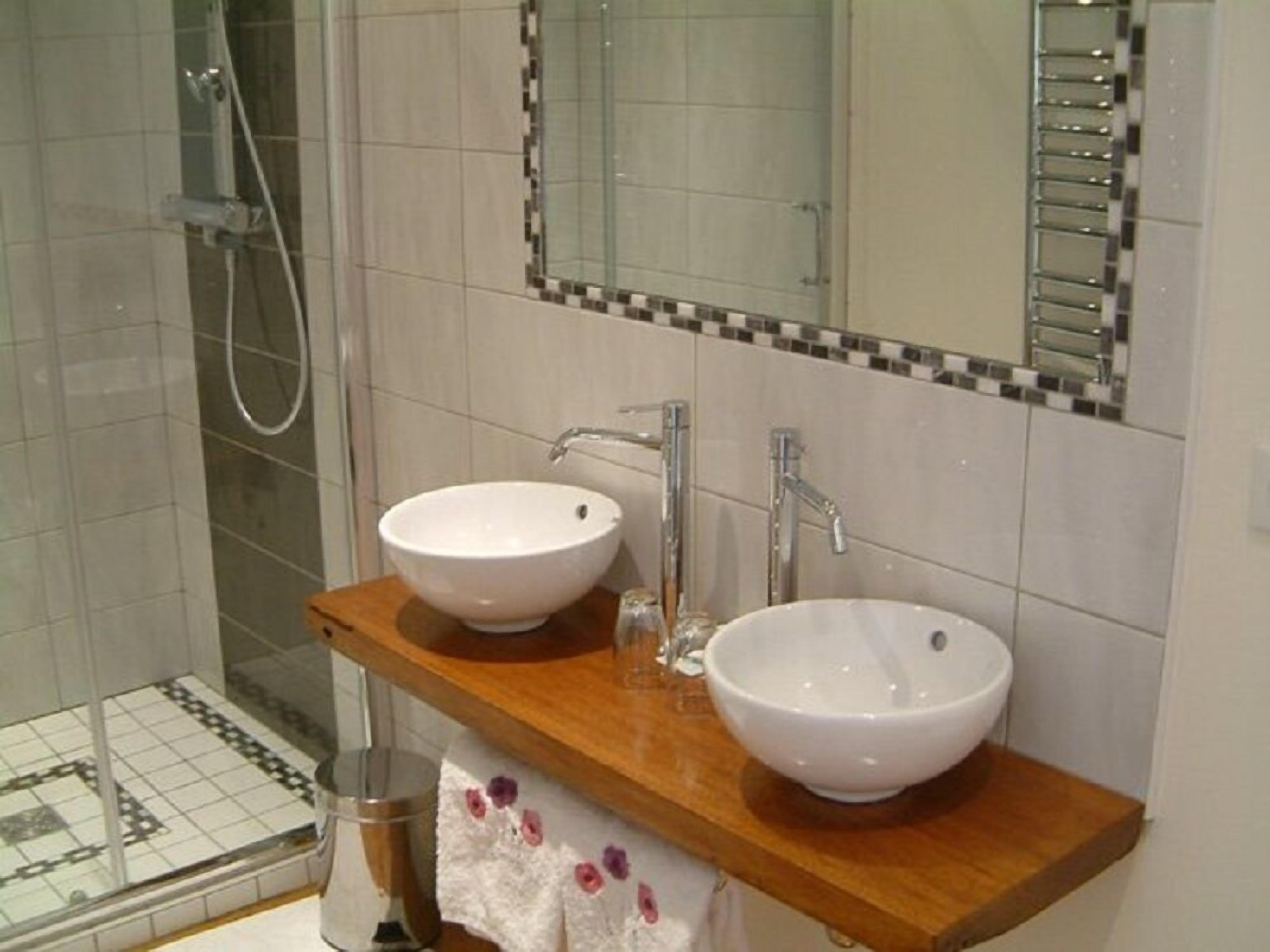 Un baño con dos lavamanos blancos, un mueble de tocador de madera, una cabina de ducha de vidrio y piso de baldosas.