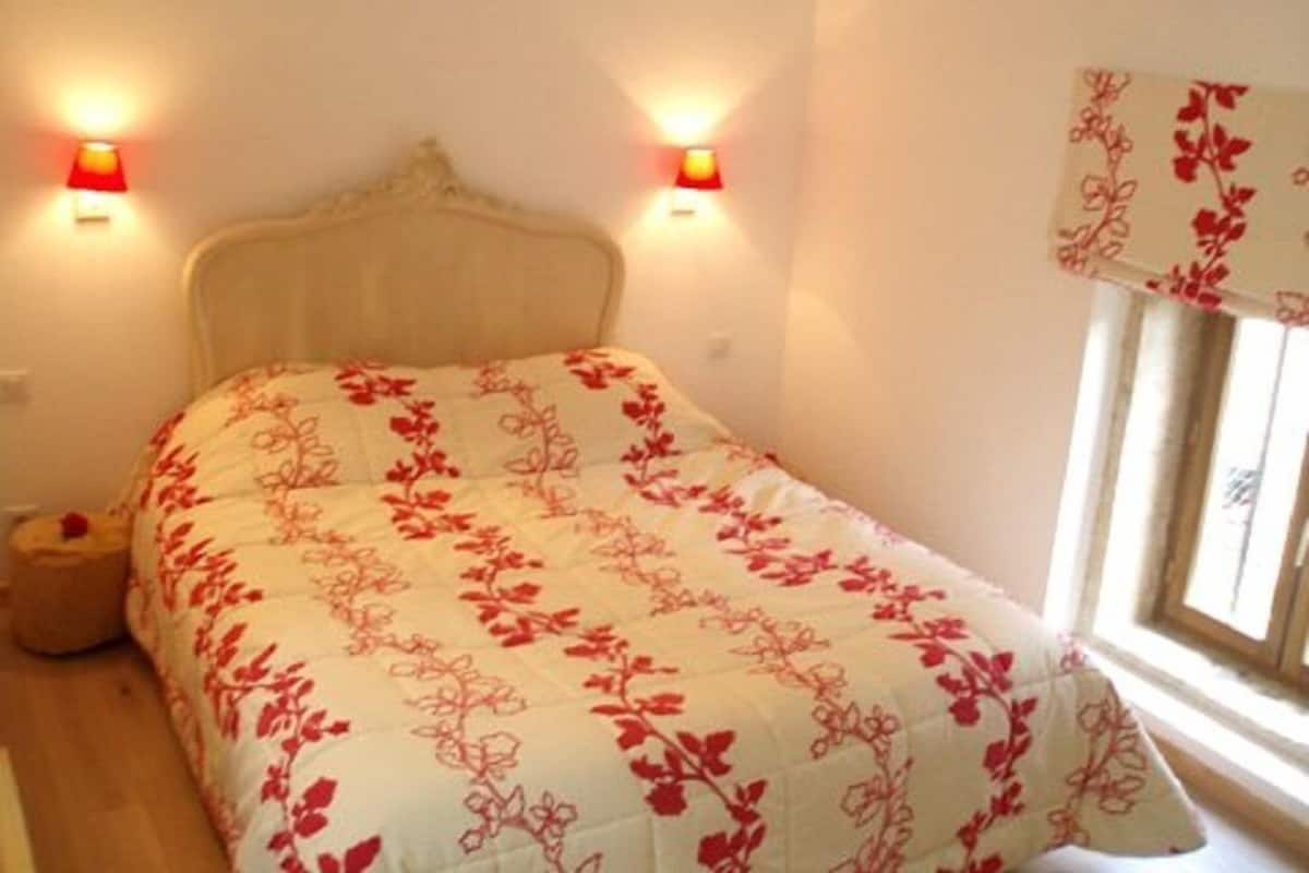 Ein Schlafzimmer mit einem Bett, zwei Wandleuchten, einem Fenster mit floralem Vorhang und einem Nachttisch.