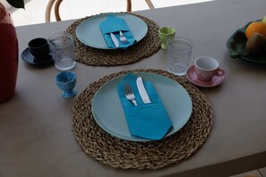 A table set with plates, cutlery, and glasses, with a woven placemat and a bowl of fruit in the background. - Maggana Residence (Kefalonia)