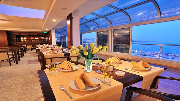 A restaurant with tables set for dining, a vase of yellow flowers, and a view of a cityscape through large windows. - Pelikan Otel (Istanbul)