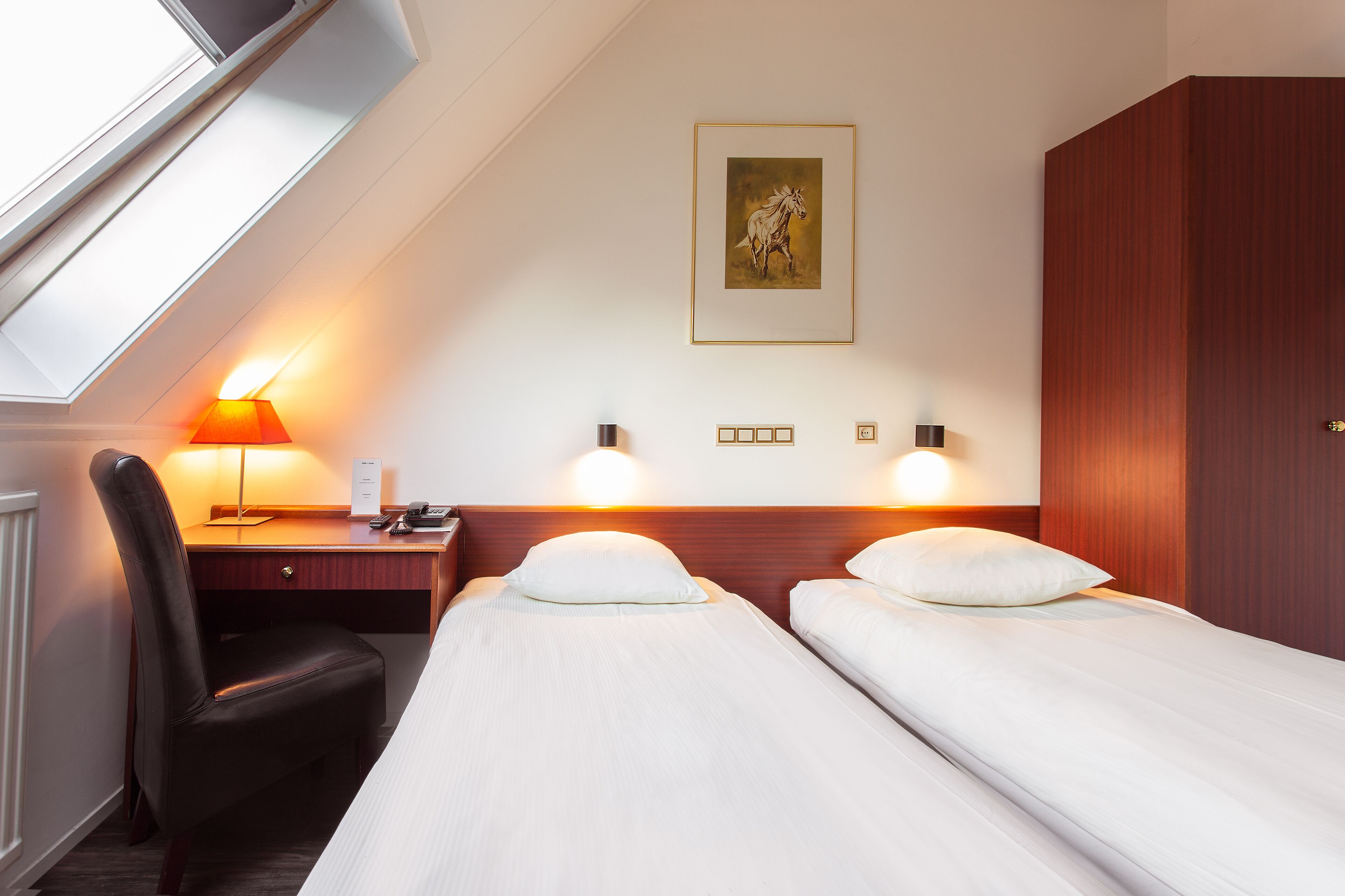 Een hotelkamer met twee enkele bedden, een bureau met stoel, een lamp en een ingelijste foto aan de muur.