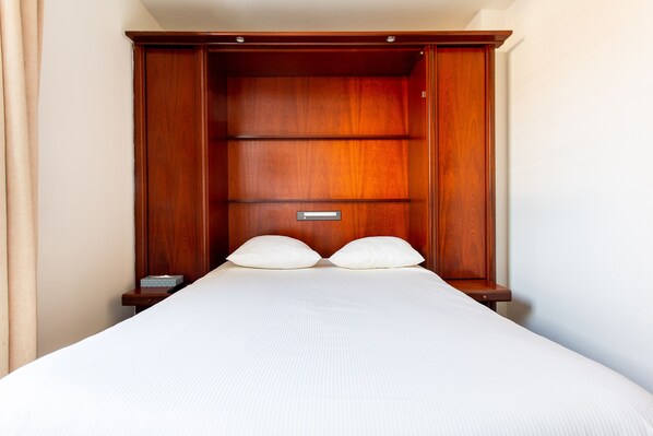 Quarto de hotel com guarda-roupa de madeira, cama com roupa de cama branca e mesa de cabeceira com abajur.