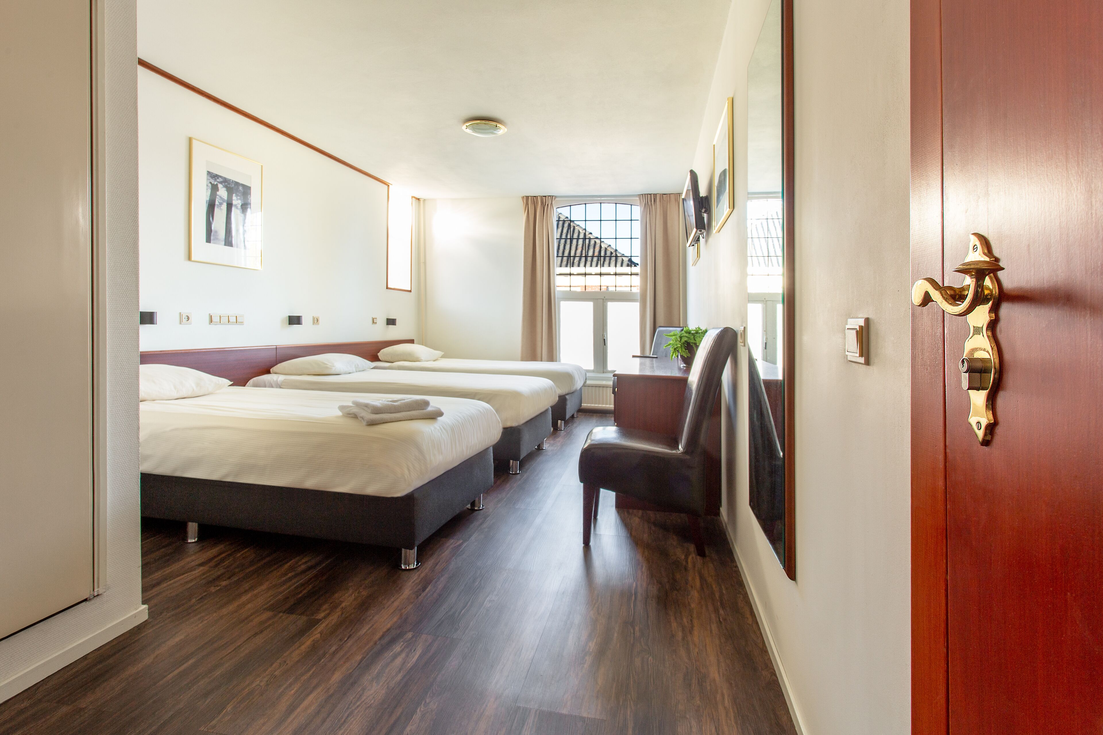 A hotel room with three beds, a wooden floor, a door with a brass handle, and a window with a view of a balcony.