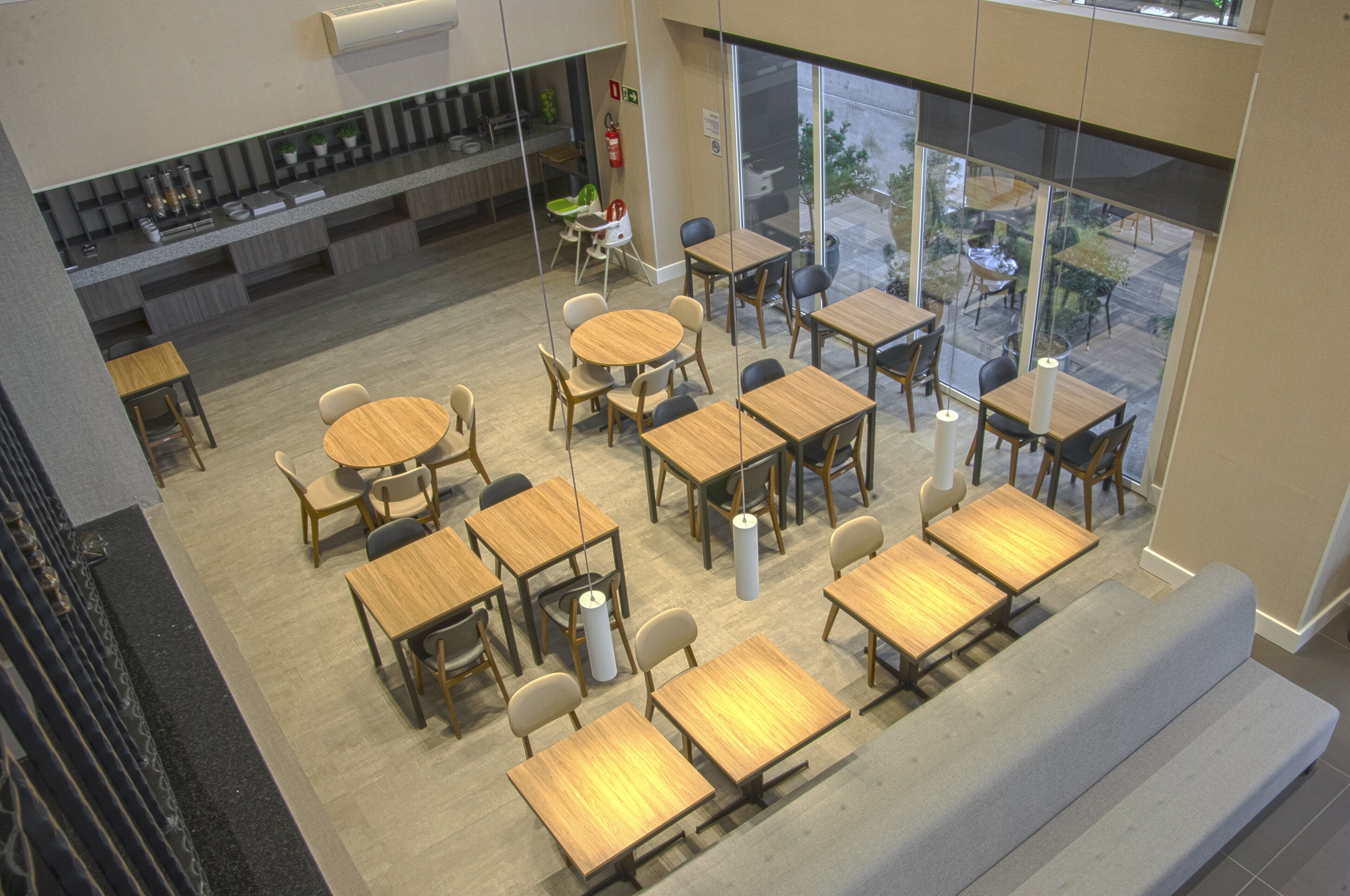 A modern cafe with multiple seating areas, including round tables and rectangular tables, and a visible outdoor view through large windows.