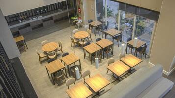 Un café moderno con varias zonas de estar, incluyendo mesas redondas y rectangulares, y una vista exterior visible a través de grandes ventanales.