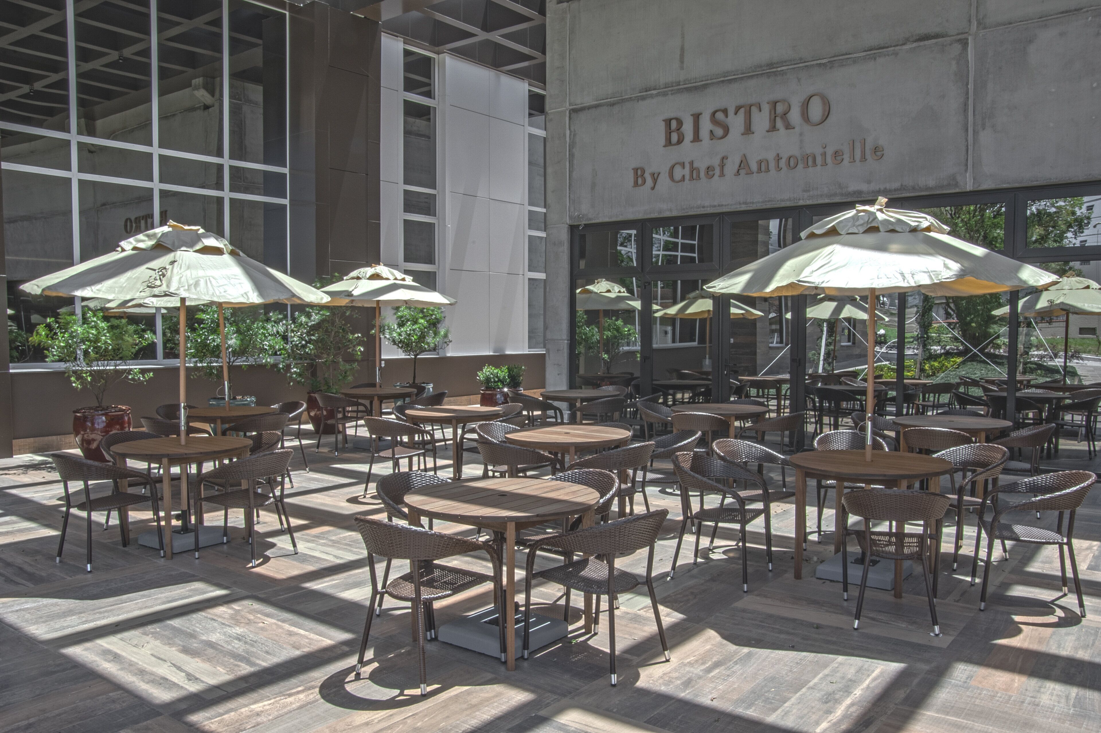 Un área de café al aire libre con mesas y sillas, sombrillas y un edificio con un letrero que dice 'BISTRO By Chef Antonielle'.
