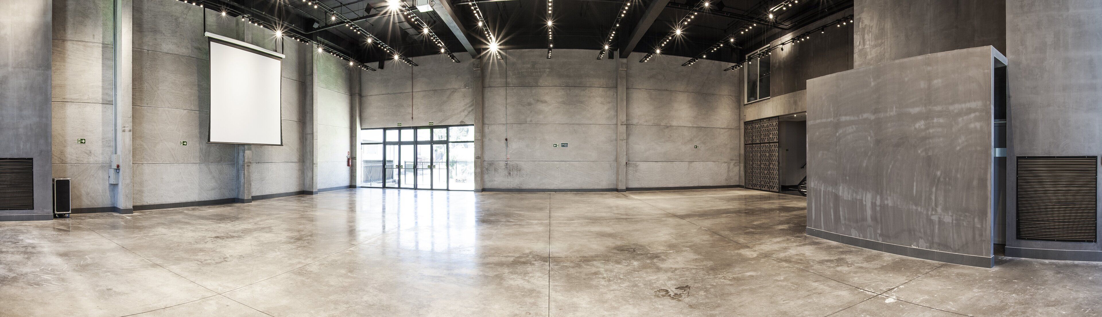 Ein großzügiger, moderner Innenraum mit hoher Decke und Designelementen im Industrielook, darunter freiliegende Balken und eine große, leere Projektionsfläche.