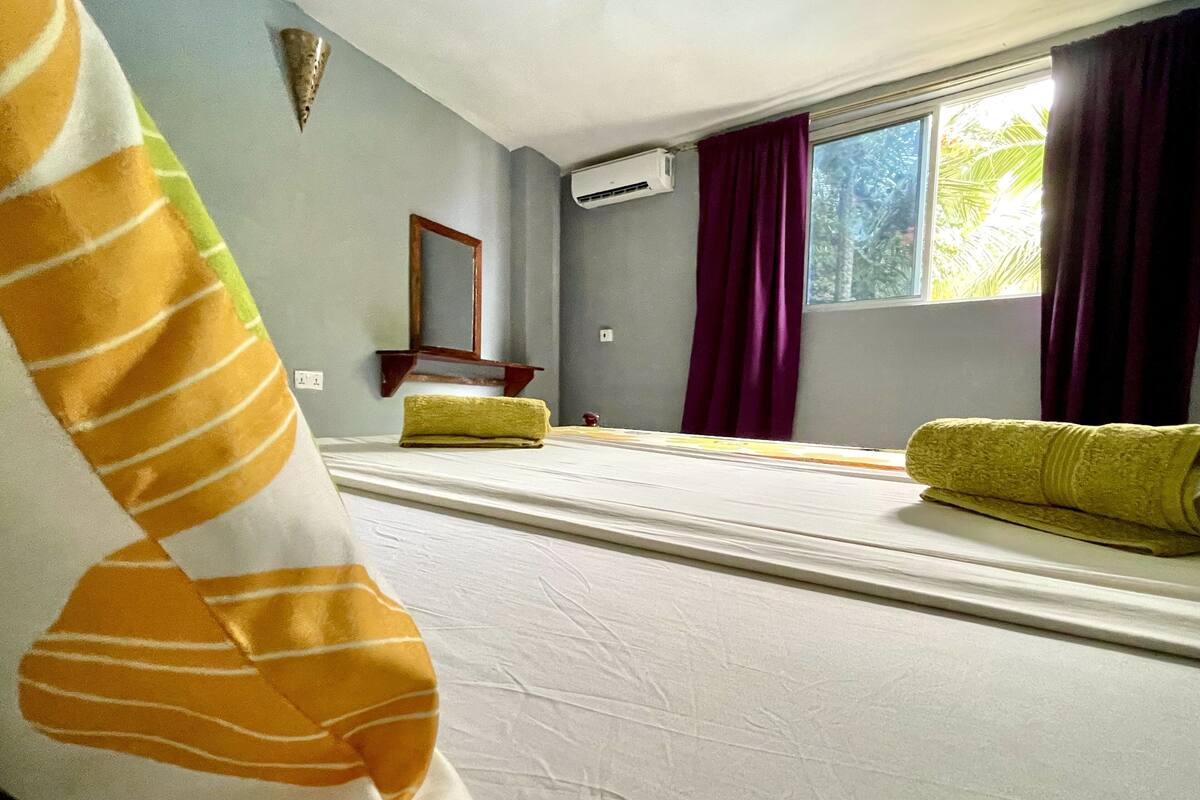 A room with a bed, a window with curtains, an air conditioning unit, and a mirror on the wall.