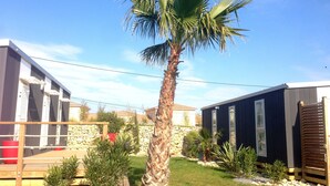 A modern outdoor area with a palm tree, a wooden deck, and two cabins with dark exteriors and white windows. - Camping Le Botanic (Fabregues)