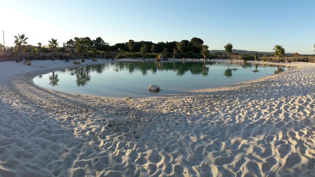 A sandy beach with a circular pool surrounded by palm trees and a clear sky. - Camping Le Botanic (Fabregues)