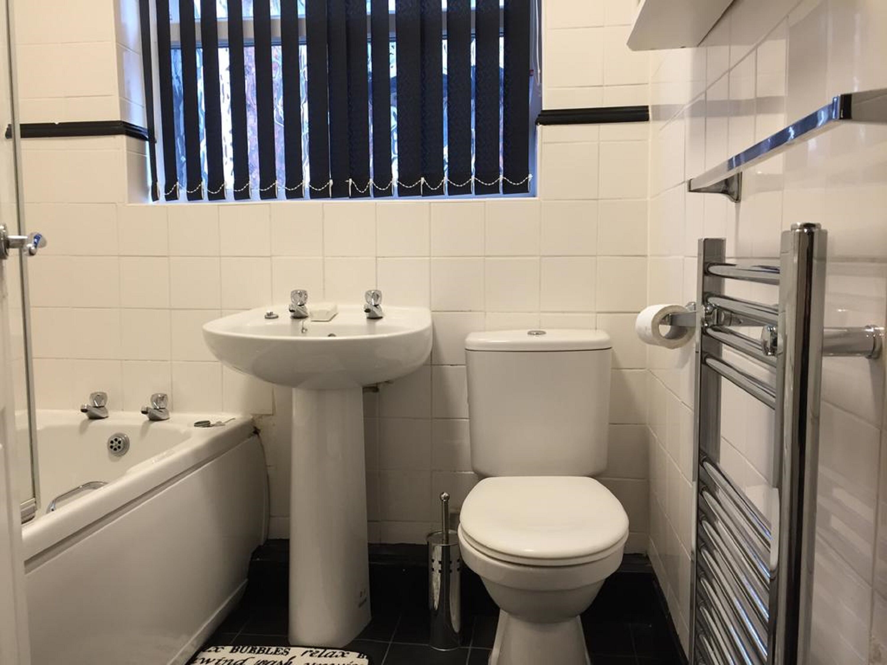 Een kleine badkamer met een wit wastafelmeubel, toilet en bad. Er is een raam met verticale zonwering en een handdoekrek is aan de rechterkant te zien.