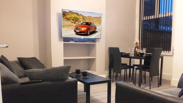 A modern living room with a sofa, a coffee table, and a TV mounted on the wall displaying a car. - Ruskin Place (Crewe)