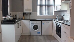 A compact kitchen with a washing machine, a built-in oven, and a window with blinds. - Ruskin Place (Crewe)
