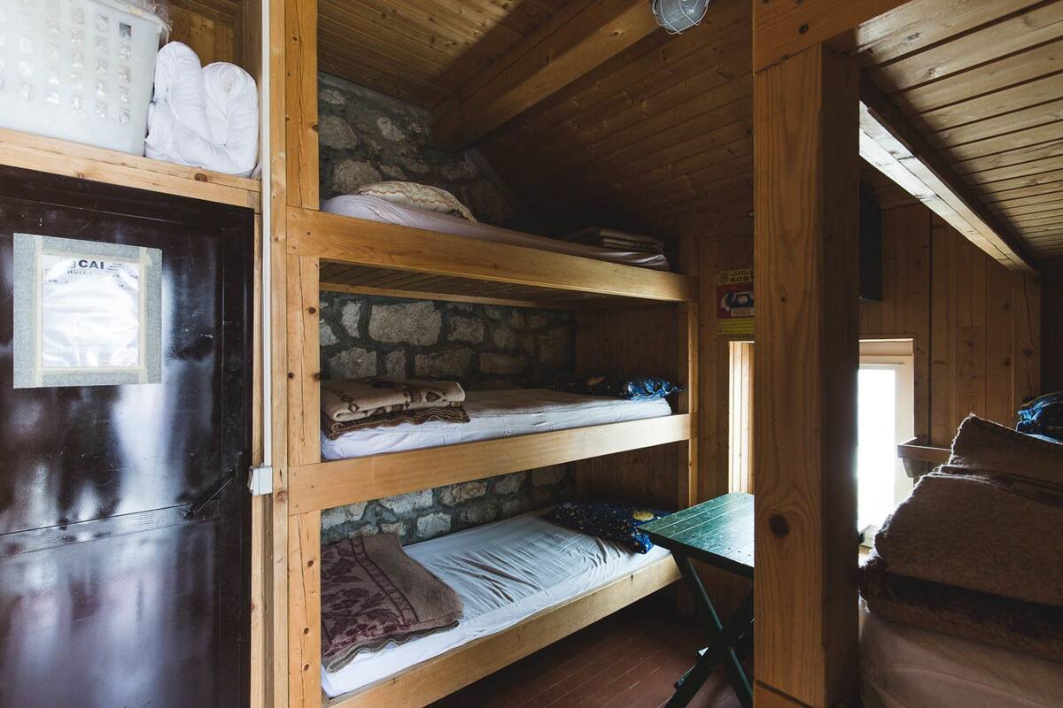 Una cabaña rústica con pared de piedra, camas individuales de madera, una puerta con un letrero y un ventanal pequeño.