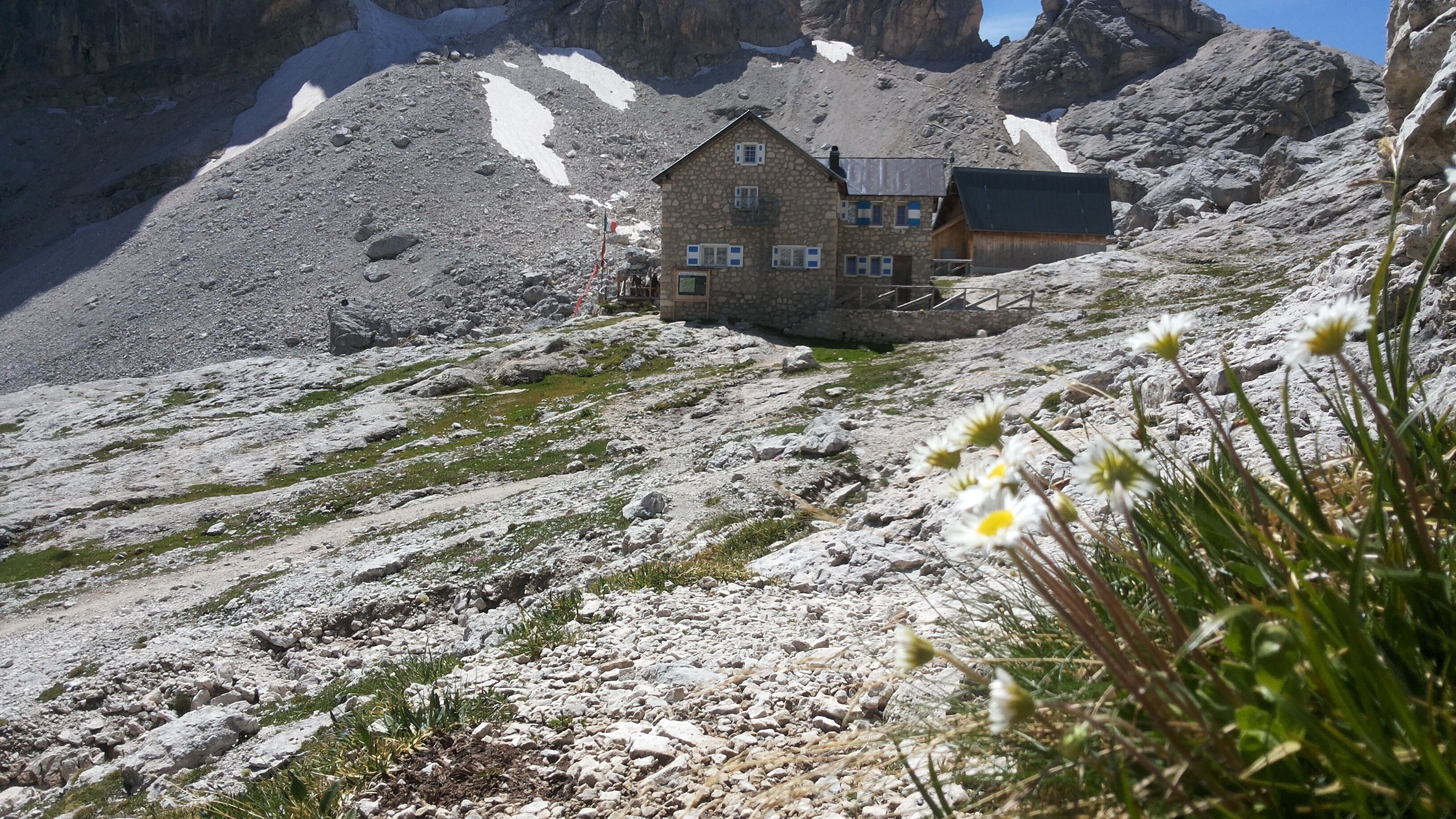 A stone building with multiple windows, situated on a rocky mountain slope with patches of snow.