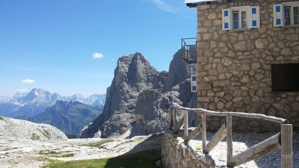 A stone building with a wooden fence, a mountain view, and a clear sky. - Rifugio Mulaz (Falcade)