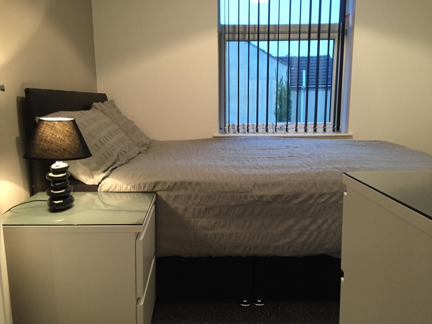 A bedroom with a bed, a bedside table with a lamp, a window with bars, and a view of buildings outside.