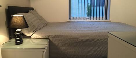 Un dormitorio con cama, mesita de noche con lámpara, ventana con rejas y vista a edificios.