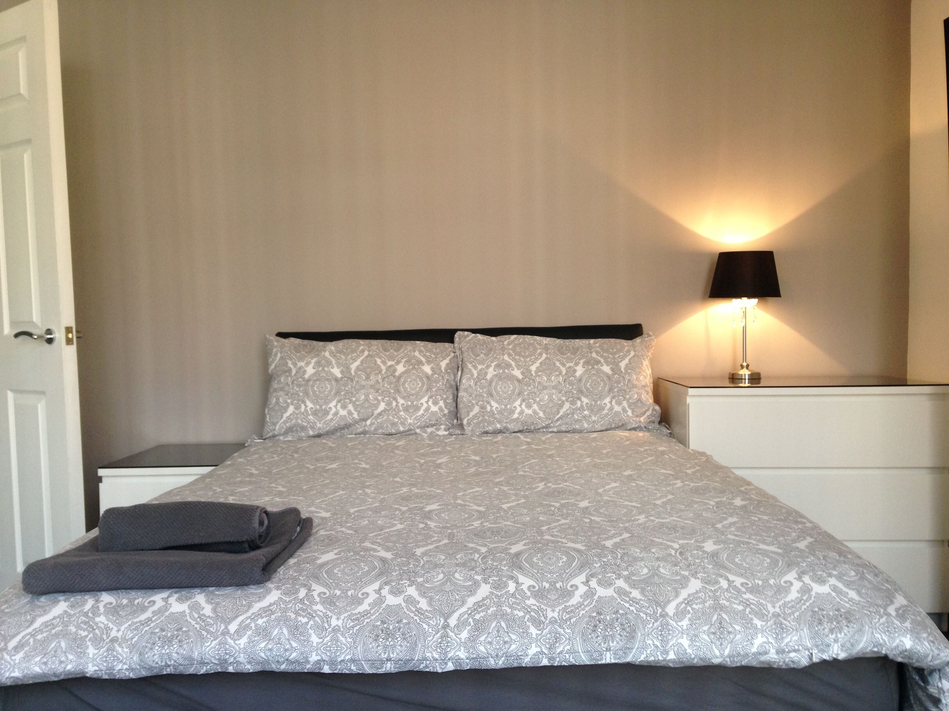 A neatly made bed with a patterned bedspread and pillows, a bedside lamp, and a folded towel on the nightstand.