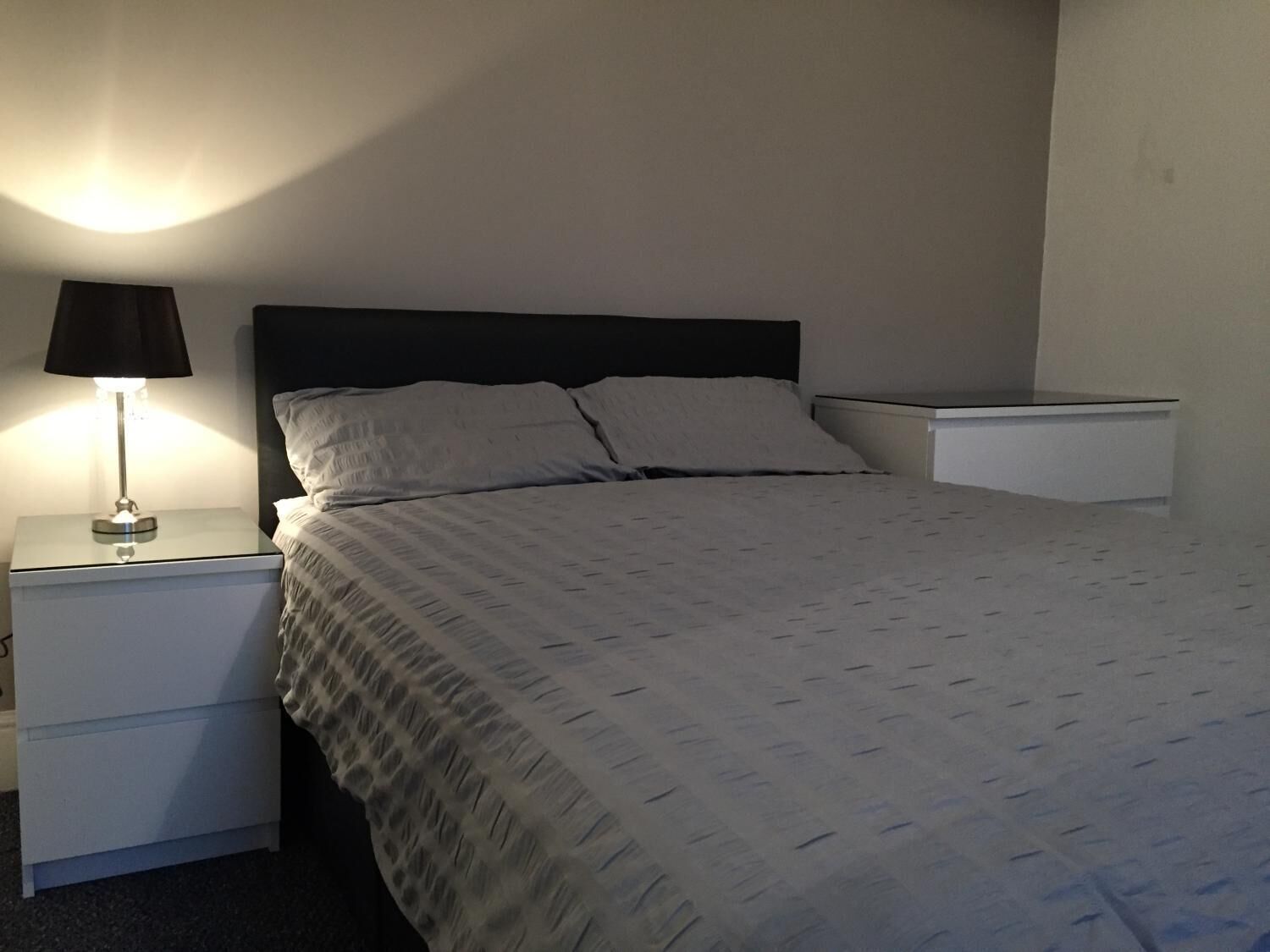 A neatly made bed with a white comforter and two pillows, a bedside table with a lamp, and a small white nightstand.