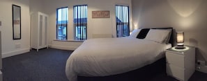 A bedroom with a large bed, a bedside table with a lamp, a wardrobe, and a window with blinds.
