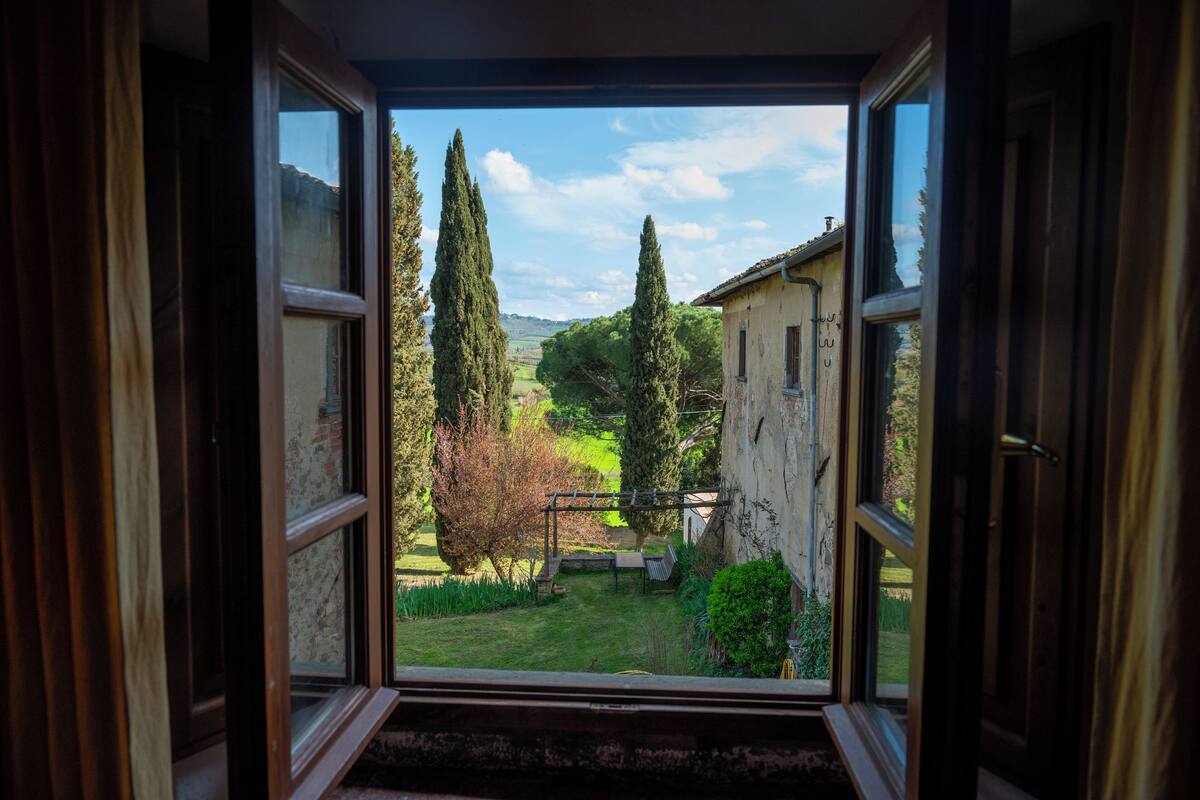 An open window revealing a garden with a pergola, trees, and a building in the background.