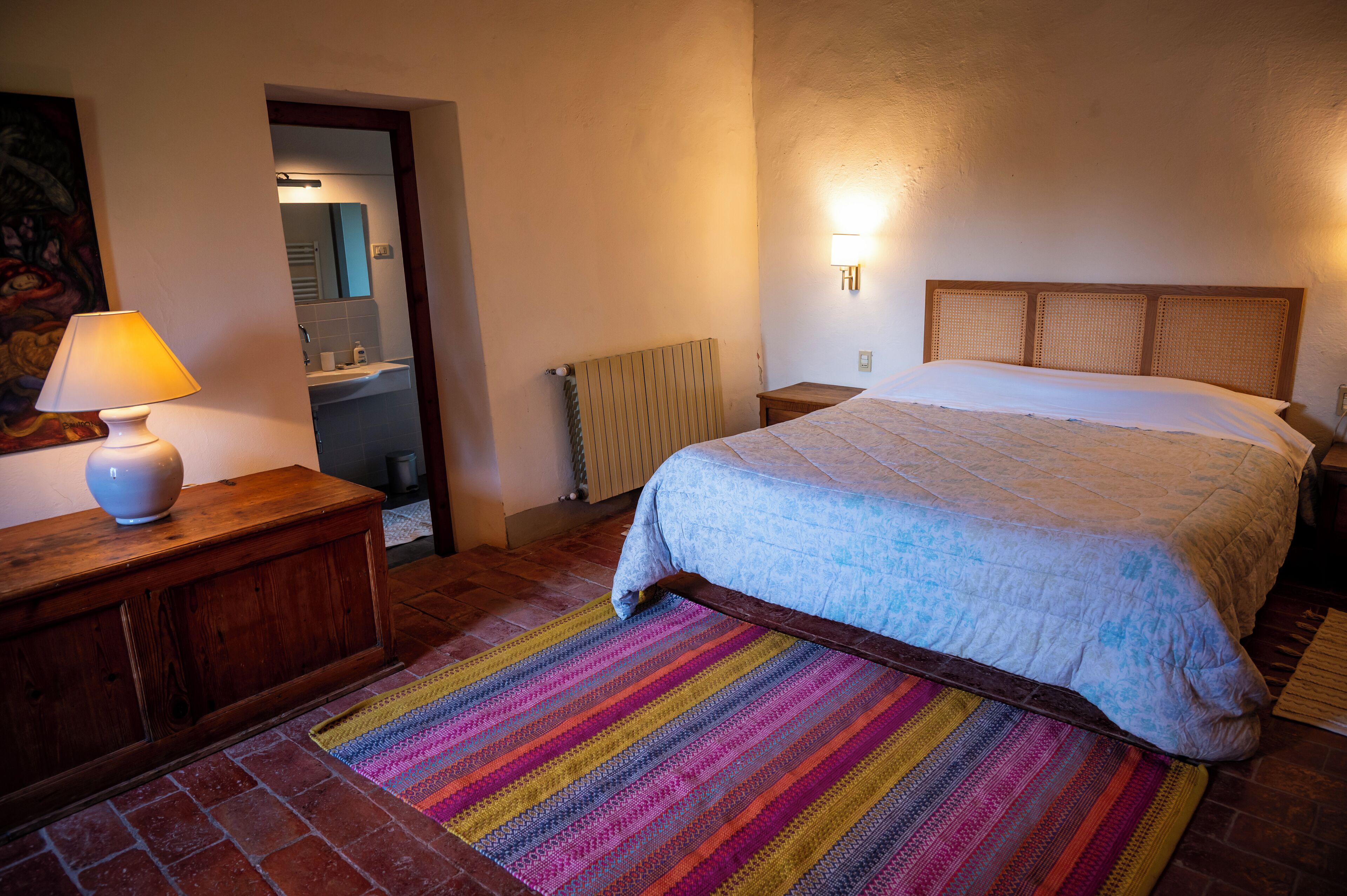 A bedroom with a bed, a nightstand, a lamp, a colorful rug, a wooden dresser, a radiator, and a bathroom visible through an open door.