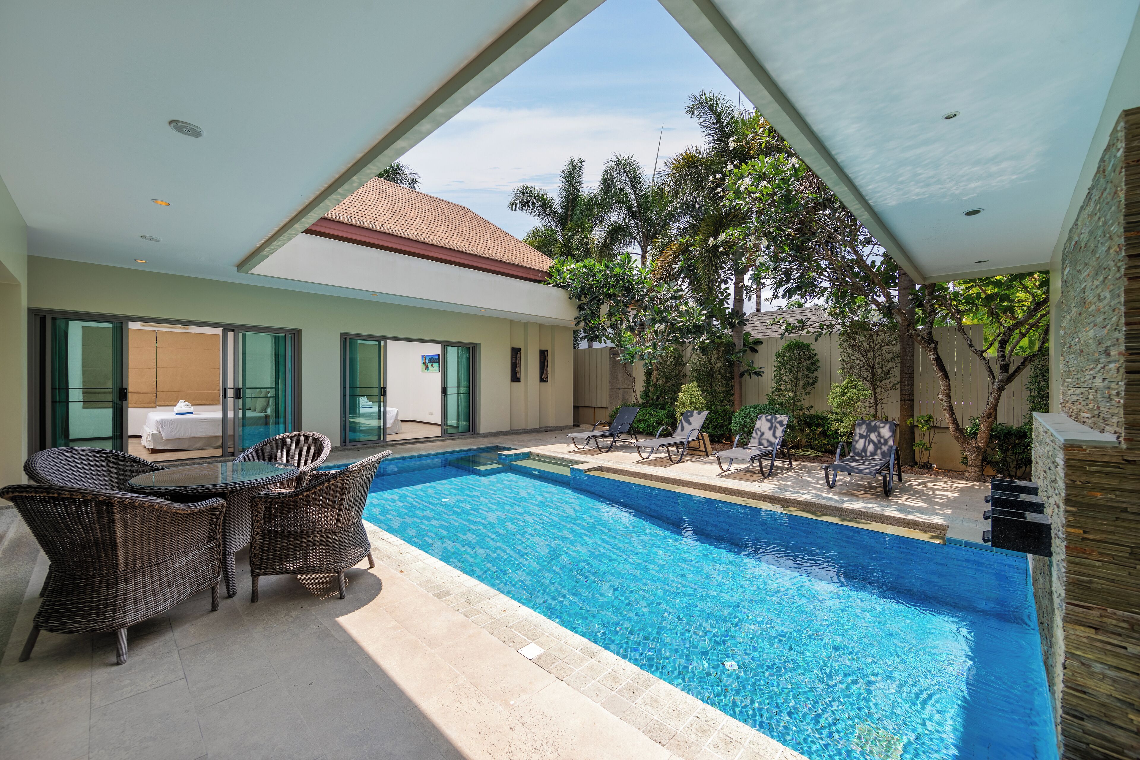 Una piscina con sillones de descanso y una mesa, rodeada de vegetación y un edificio con tejado de baldosas.