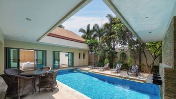 A swimming pool with lounge chairs and a table, surrounded by greenery and a building with a tiled roof.