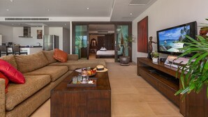 A modern living room with a sofa, coffee table, and TV. There is a kitchen in the background and a bedroom with a bed and a plant. - Serenity Grand Seaview Suite (Rawai)