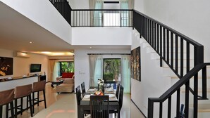 Un vaste espace de vie comprenant une salle à manger avec une table dressée pour six personnes, un coin cuisine équipé d’un téléviseur et un escalier menant à l’étage supérieur.