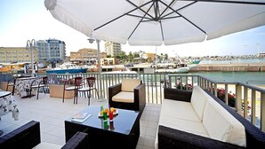 Una terraza con mobiliario exterior, vista al puerto deportivo y edificios al fondo.