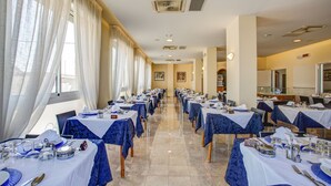 Un salón de banquetes con filas de mesas dispuestas para cenar, amplias ventanas con cortinas y una cocina visible al fondo.