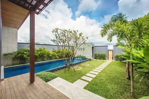 A modern outdoor pool area with a wooden deck, a stone wall, and a variety of tropical plants.