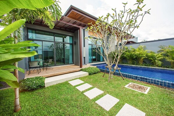 A modern house with a glass door entrance, a wooden deck, a small pool, and a well-maintained lawn.