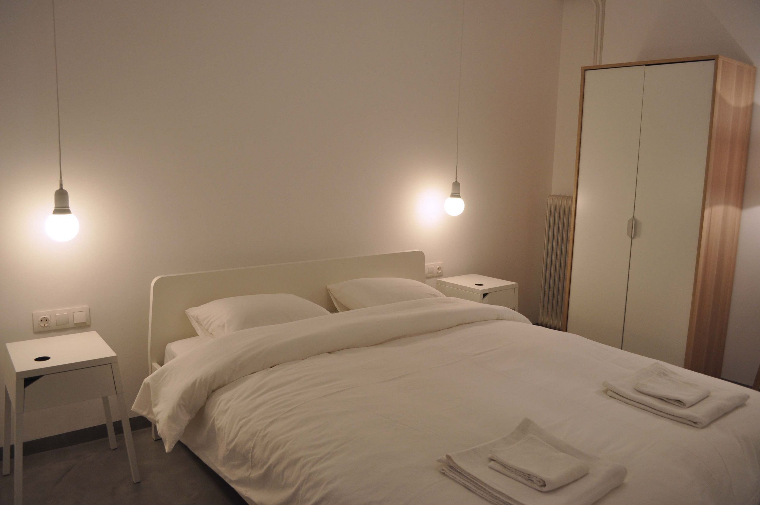 Una cama bien hecha con ropa blanca, una mesita de noche con lámpara y un armario en una habitación de hotel.