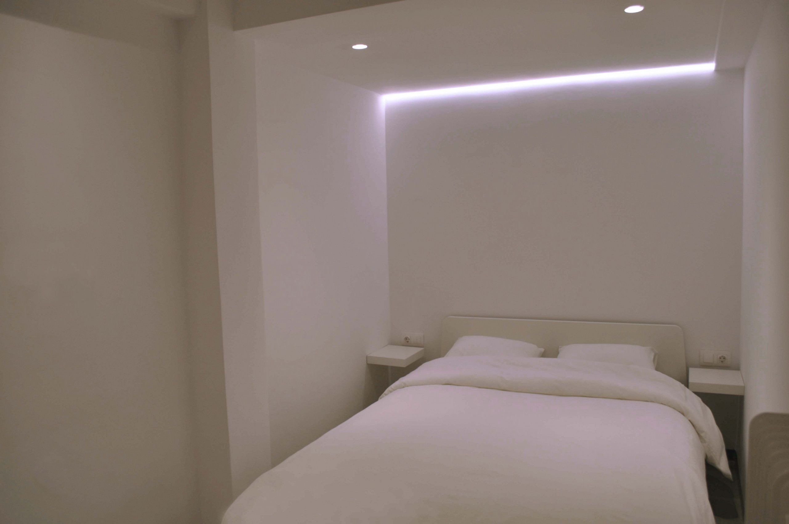 Quarto minimalista com uma cama de solteiro, mesas de cabeceira e iluminação embutida no teto.