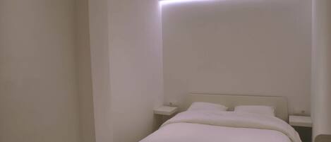Quarto minimalista com uma cama de solteiro, mesas de cabeceira e iluminação embutida no teto.
