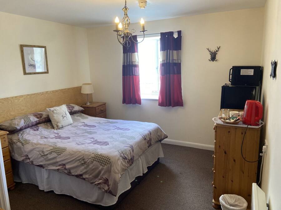A bedroom with a bed, nightstand, lamp, curtains, a chandelier, a window, a wall-mounted heater, a wooden dresser, a trash bin, and a wall-mounted TV.