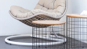 Una silla colgante con asiento y respaldo acolchados, suspendida por un marco metálico curvo, ubicada junto a una mesita redonda con base de alambre.