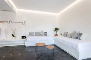 A modern, minimalist bedroom with a white bed, two round wooden tables, a potted plant, and a wall-mounted TV. - Aegean Gem (Santorini)