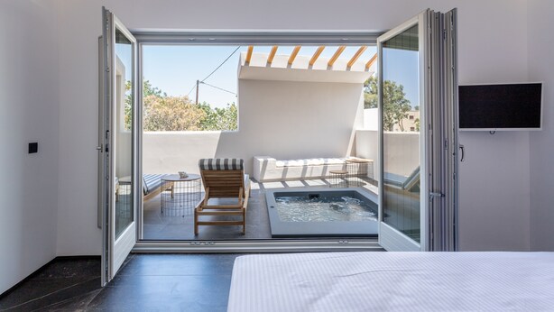 Una habitación de hotel moderna con un gran ventanal corredero que da a una zona de estar exterior, un jacuzzi pequeño y una cama con edredón blanco.