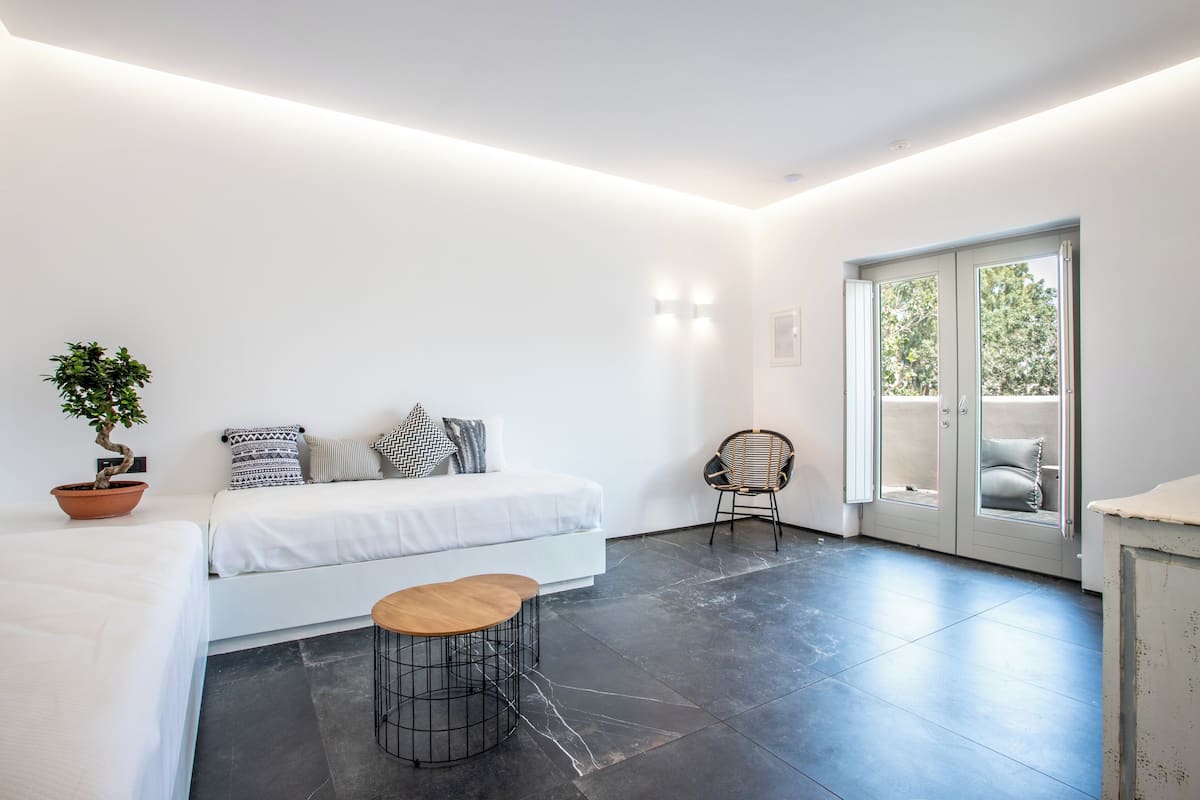 A modern room with a bed, a chair, a small table, a potted plant, and a sliding glass door leading to an outdoor area.