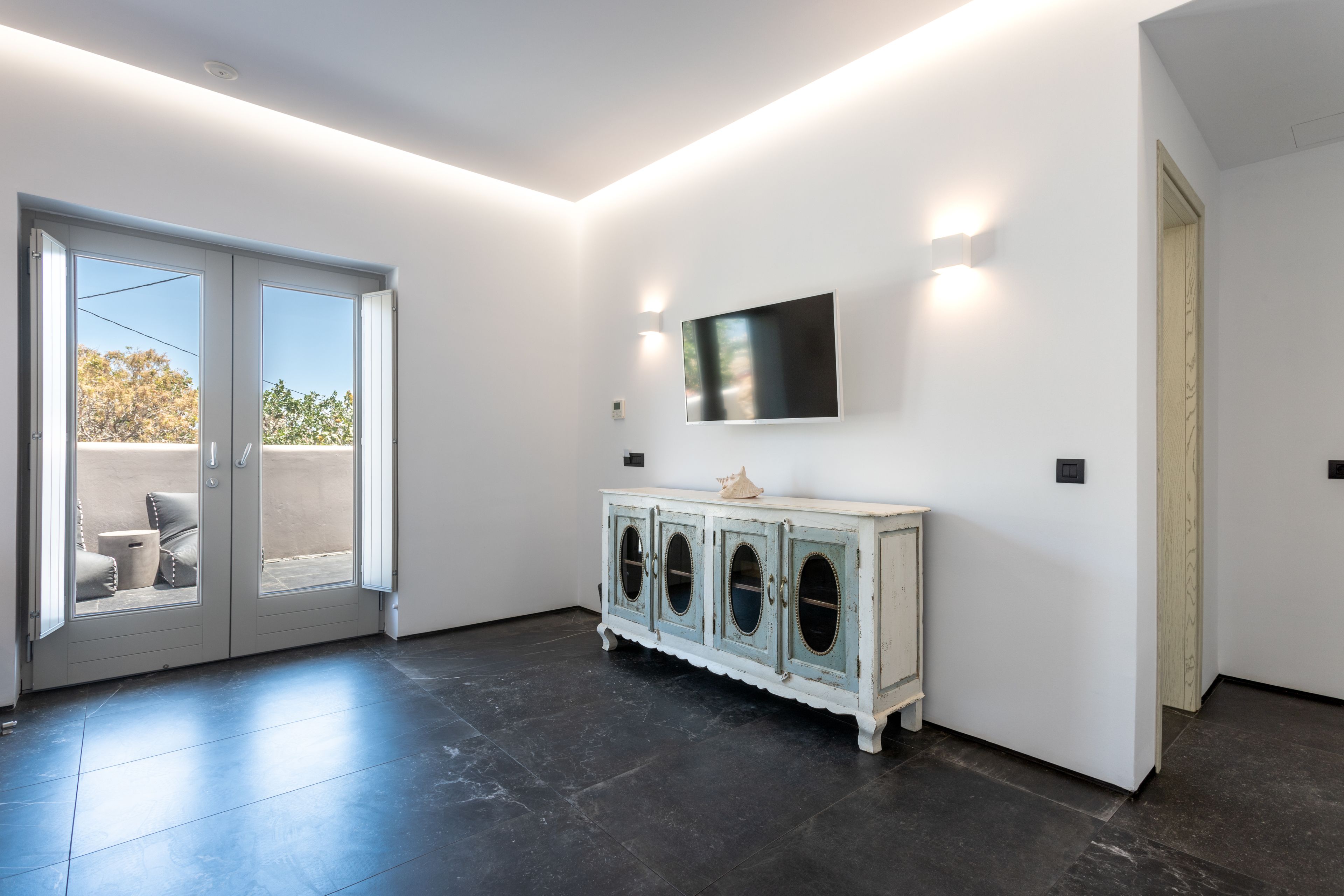 A modern room with a large sliding glass door leading to an outdoor area, a white cabinet with a flat-screen TV mounted above it, and a dark tiled floor.