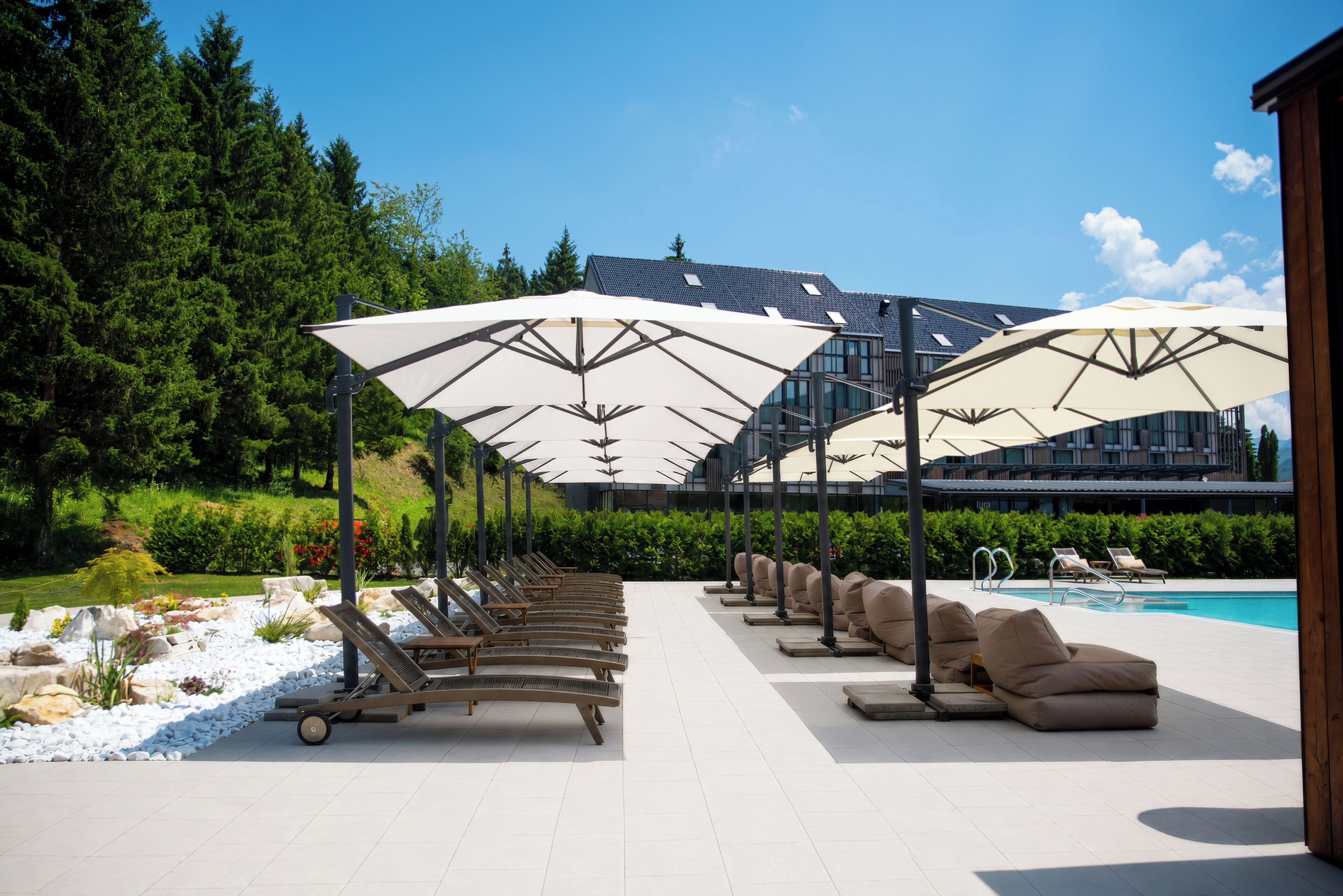 A poolside area with sun loungers and umbrellas, a pool, and a building with a sloped roof in the background.